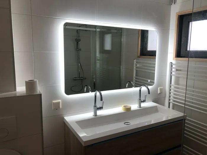 Badkamer met verlichte LED-spiegel boven dubbele wastafel, inloopdouche zichtbaar in de spiegel