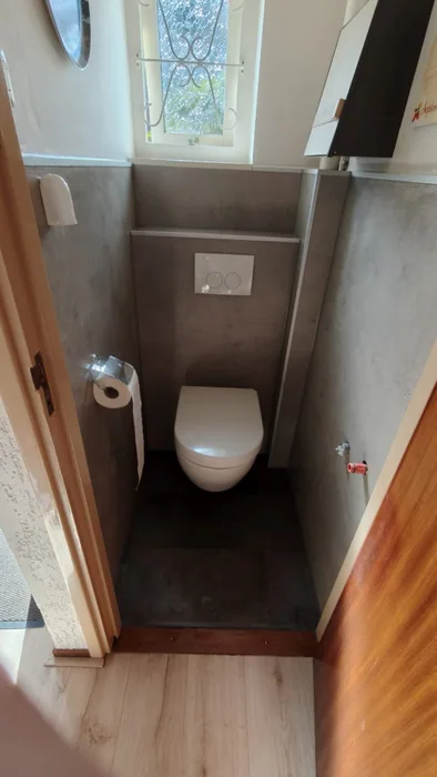 Gerenoveerd toilet met hangtoilet, grijze betonlook wandpanelen, voorzetwand met bedieningsplaat en raampje met sierrooster