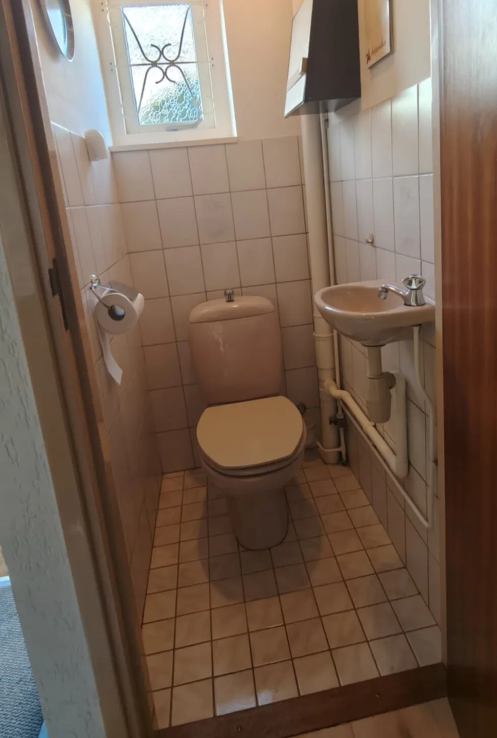 Oud toilet met beige staand closet, klein fonteintje, witte tegels en raampje met sierrooster
